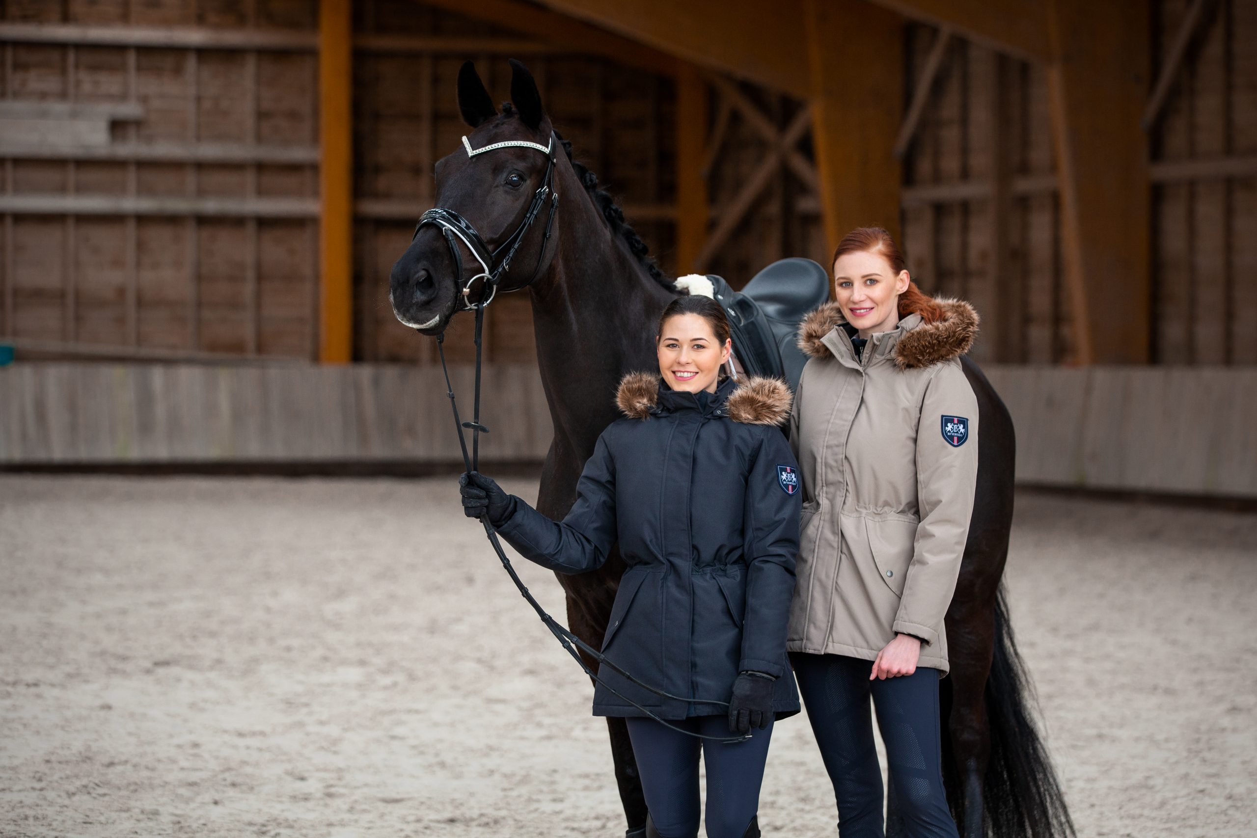 two riders in winter jackets standing in front of a black horse in indoor arena
